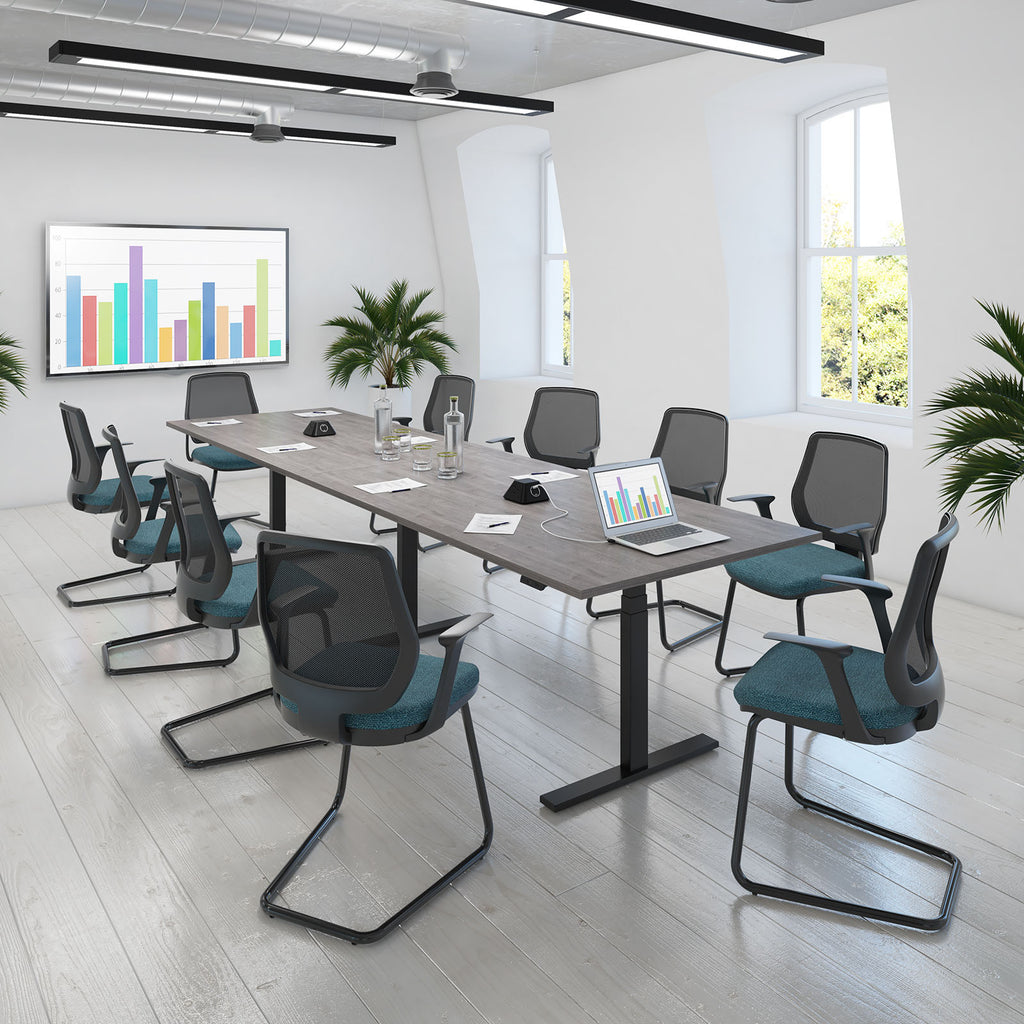 A modern conference room setup with a long rectangular desk and multiple mesh office chairs featuring dark gray frames and teal-blue seat cushions, placed in a bright white room with large windows and a framed multicolor bar chart on the wall.
