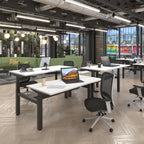 Modern open-plan office with multiple white desks, black metal frames, mesh office chairs, laptops, and a bright green sofa in the background.