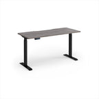 A modern height-adjustable desk with a grey wood-grain top and black metal legs, shown on a clean white background.