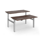 Two back-to-back height adjustable desks with shared silver metal frame and wood-effect tops on a white background