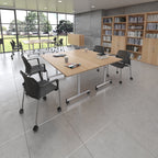 A modern office meeting room with a light wood rectangular conference table on casters, surrounded by black mesh office chairs with silver metal legs. Large windows on the left look out onto a green field, and light wood bookshelves line the right wall.