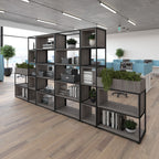 A large modular open shelving unit with dark grey wood-grain panels and black metal framing, styled with books, plants, and storage boxes in a modern office environment.