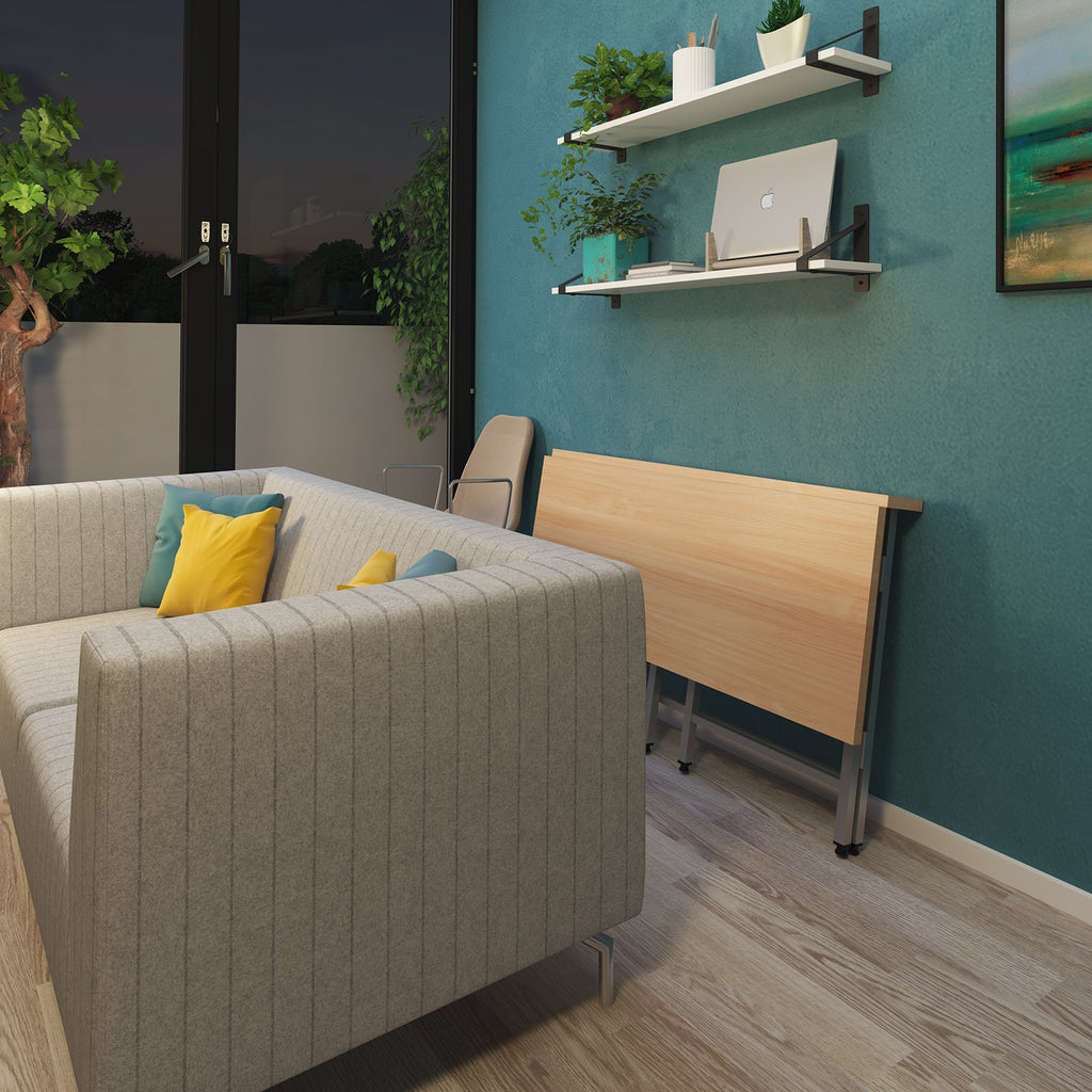 A light wood folding table with metal legs is shown in a modern room setting, folded against a teal wall next to a grey ribbed sofa with yellow and blue pillows. The wall features two white floating shelves with small potted plants and a laptop.