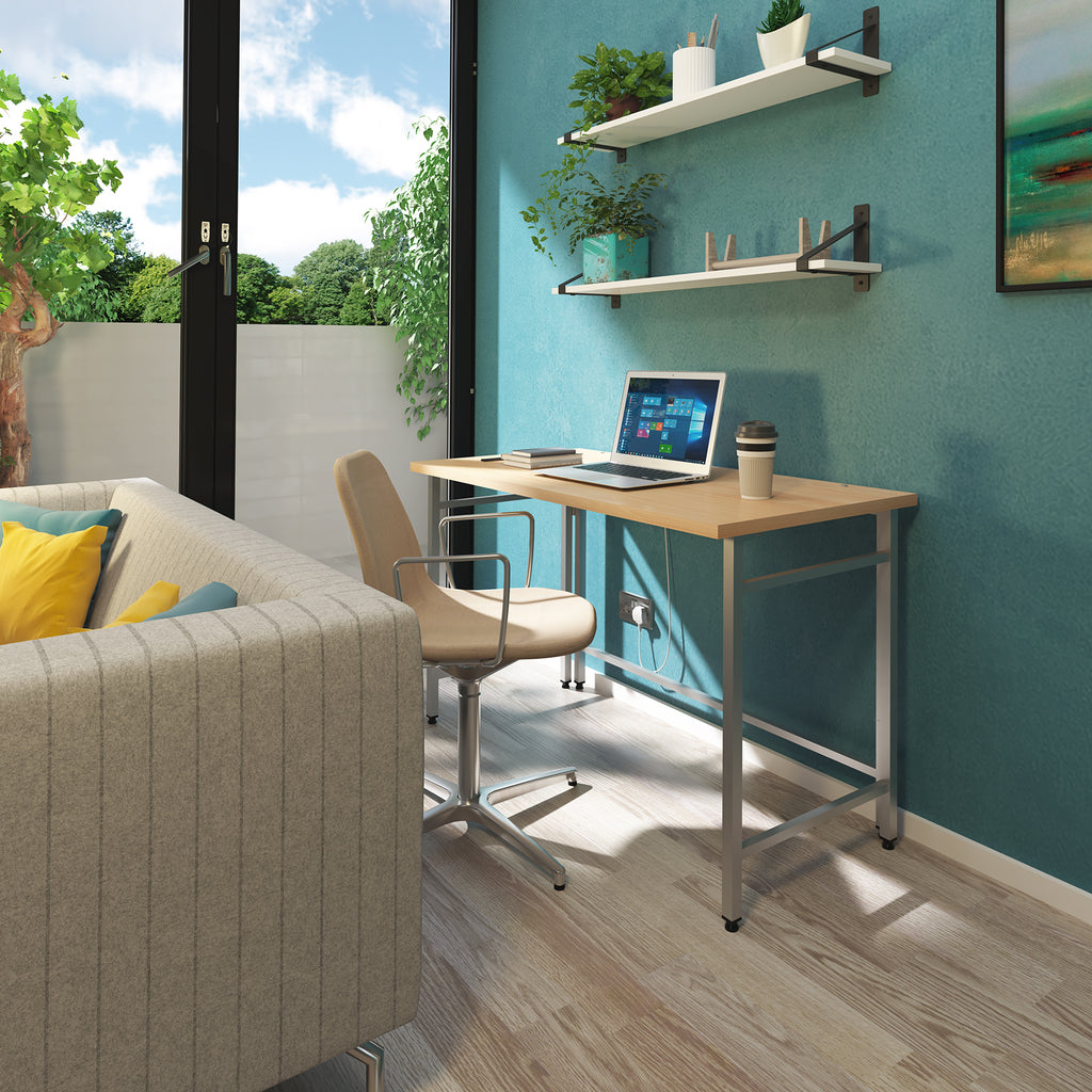A modern home office setup featuring a light wood desk with a silver metal frame, paired with a beige swivel office chair. A laptop sits on the desk, and the setup is mounted against a teal wall with floating shelves and plants.