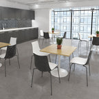 A modern dining or meeting space with multiple chairs and tables arranged in a bright interior. The room includes dark grey cabinets, a tiled backsplash, large floor-to-ceiling windows, and grey tile flooring.