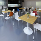 A modern office or cafeteria interior with multiple light wood square tables and white chairs, some with coloured seat backs in orange, blue, red, and black. The room has grey flooring, dark cabinetry along the back wall, and a few decorative flowers on the tabletops.