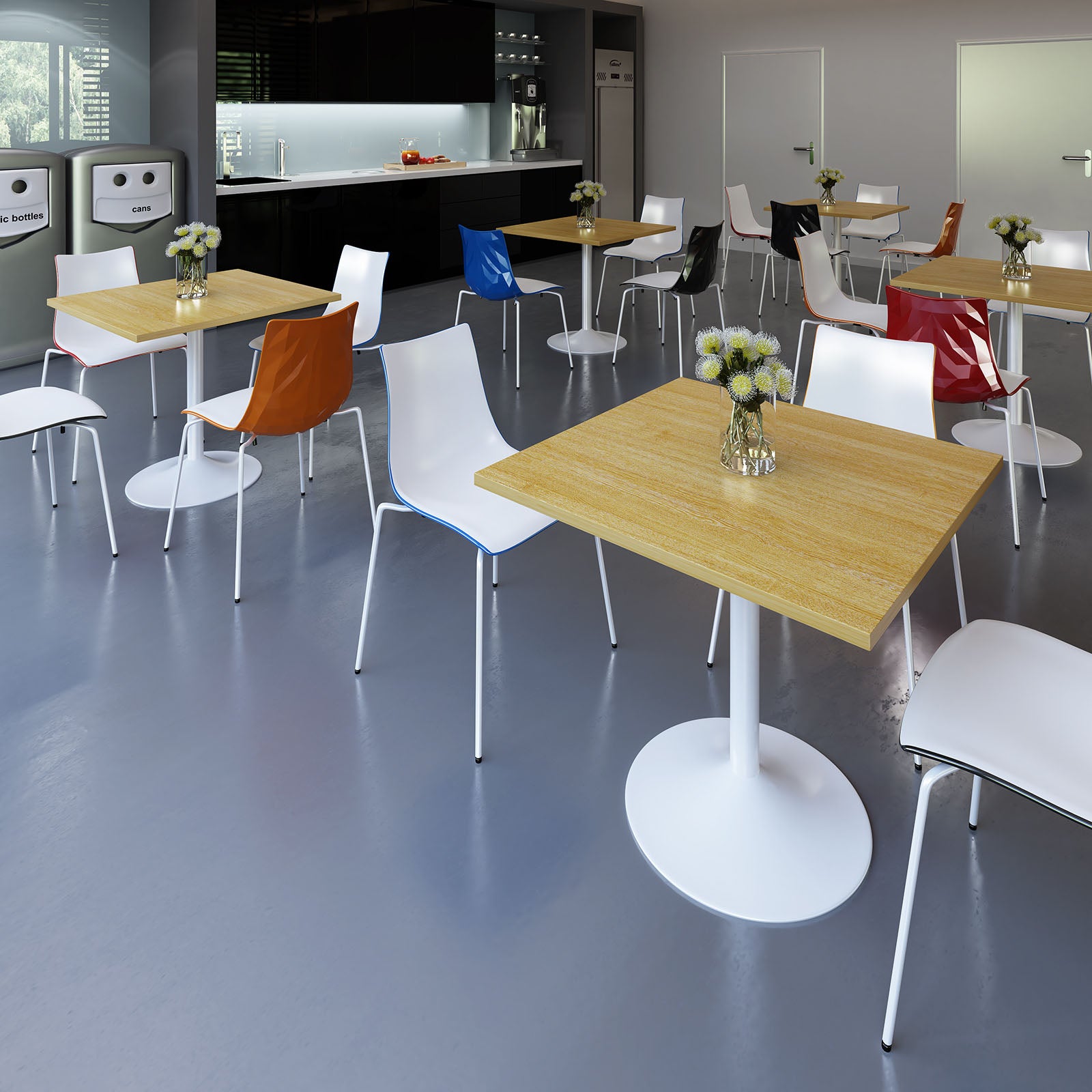 A modern office or cafeteria interior with multiple light wood square tables and white chairs, some with coloured seat backs in orange, blue, red, and black. The room has grey flooring, dark cabinetry along the back wall, and a few decorative flowers on the tabletops.
