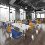 A modern office or café interior with white rectangular tables and chairs in blue, orange, and yellow, set on dark wood flooring with large windows showing a city skyline.