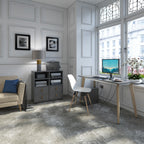 A modern home office interior featuring a grey textured rug, a white desk with a computer setup, a grey storage cabinet, and a beige armchair with a blue pillow, set against white panelled walls.