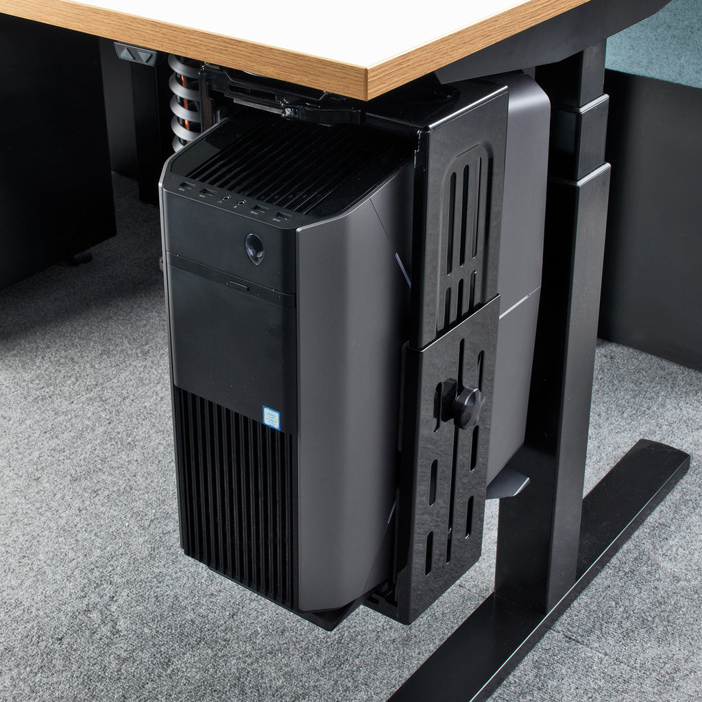 A black under-desk computer tower mounted on a black adjustable desk support bracket, shown tucked beneath a light wood-topped desk on a grey carpeted floor.