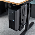 A black under-desk computer tower mounted on a black adjustable desk support bracket, shown tucked beneath a light wood-topped desk on a grey carpeted floor.