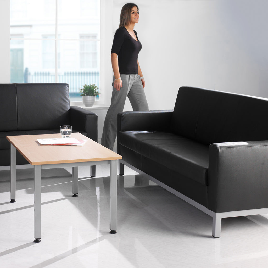 Modern office waiting area with black leather couches, a wooden coffee table, and a woman standing in the background.