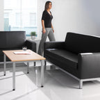 Modern office waiting area with black leather couches, a wooden coffee table, and a woman standing in the background.