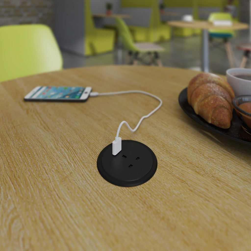 A round black table outlet socket on a light wood table, with a white charging cable plugged in and a smartphone nearby in a café setting.
