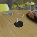 A round black table outlet socket on a light wood table, with a white charging cable plugged in and a smartphone nearby in a café setting.