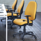 A row of modern office chairs with bright yellow upholstered seats and backs and black bases, armrests, and headrest supports, arranged in a room with dark wood flooring and a white desk on the left.