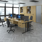 Modern open-plan office with wooden desks, ergonomic chairs, desktop computers, storage cabinets, and large windows letting in natural light.