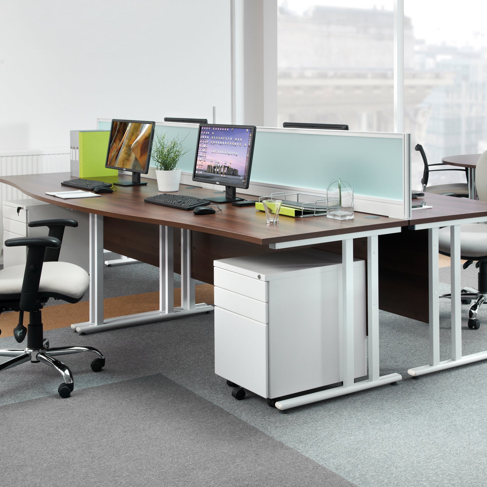 Modern office desk setup with multiple monitors, chairs, and a filing cabinet.