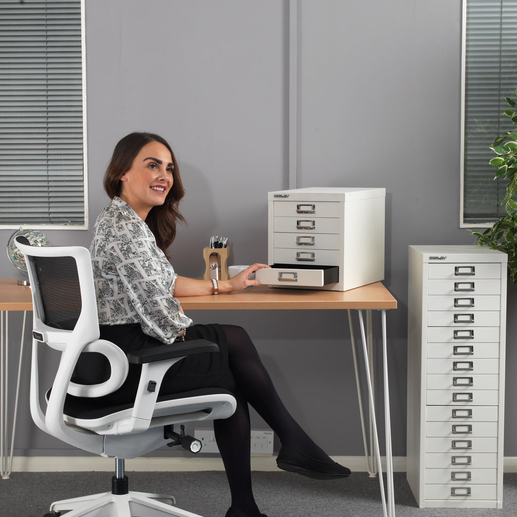 A woman sits in a white mesh office chair at a light wood desk, interacting with a white multi-drawer filing cabinet. Another matching tall filing cabinet stands on the right against a grey wall in an office setting.