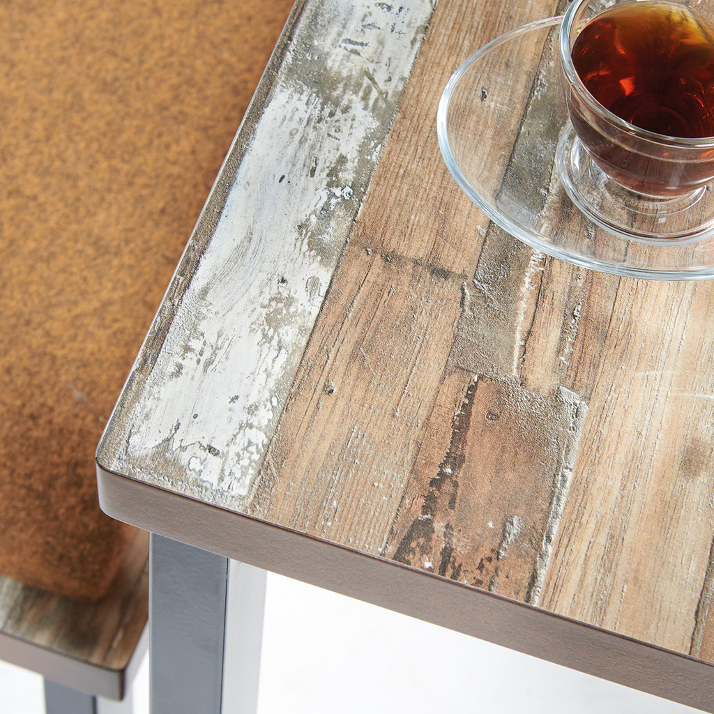 Close-up of an industrial-style table showing a distressed reclaimed wood-effect tabletop with metal accents, alongside a glass cup and cushioned bench seat.