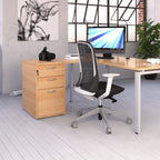 Office desk with a light wood-effect top, white metal legs, and a matching three-drawer pedestal, paired with a mesh-backed task chair, shown in a modern office setting.