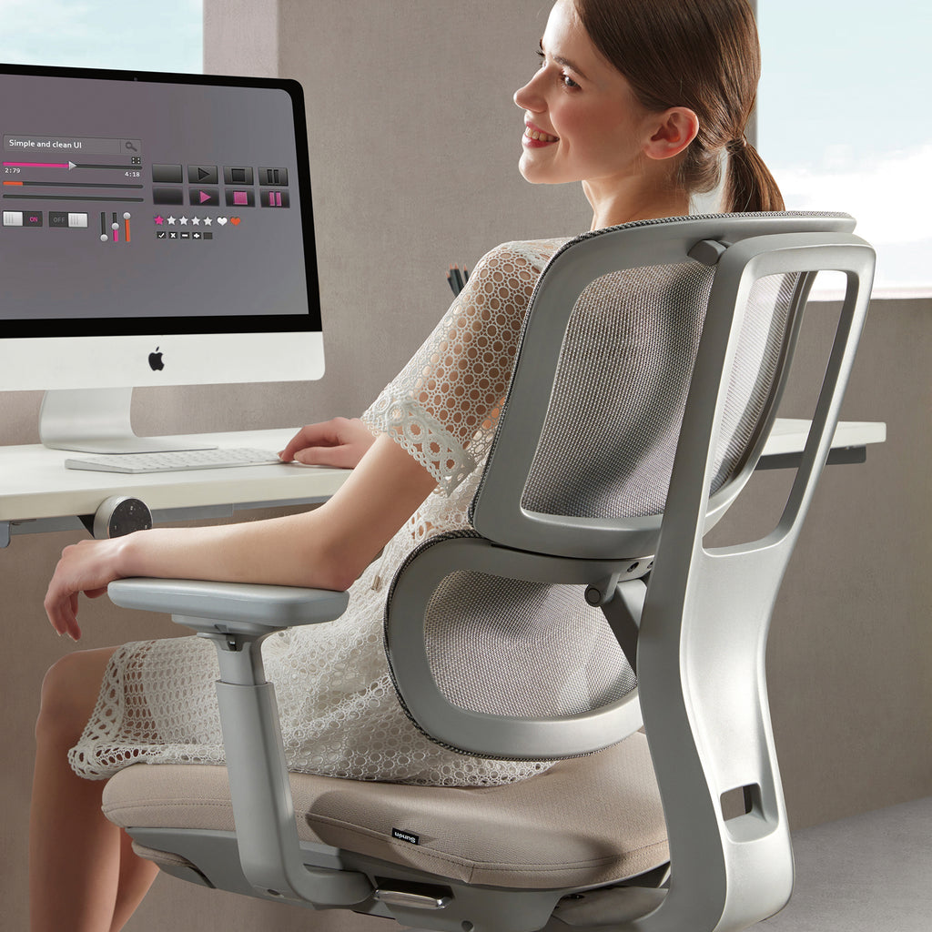 Woman seated at a desk using a computer, showcasing a grey ergonomic office chair with mesh backrest and adjustable support in a modern workspace.