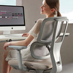Woman seated at a desk using a computer, showcasing a grey ergonomic office chair with mesh backrest and adjustable support in a modern workspace.