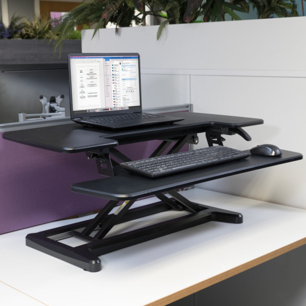 Height-adjustable desk converter with laptop and keyboard tray on an office workstation, supporting sit-stand working.