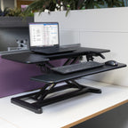 Height-adjustable desk converter with laptop and keyboard tray on an office workstation, supporting sit-stand working.