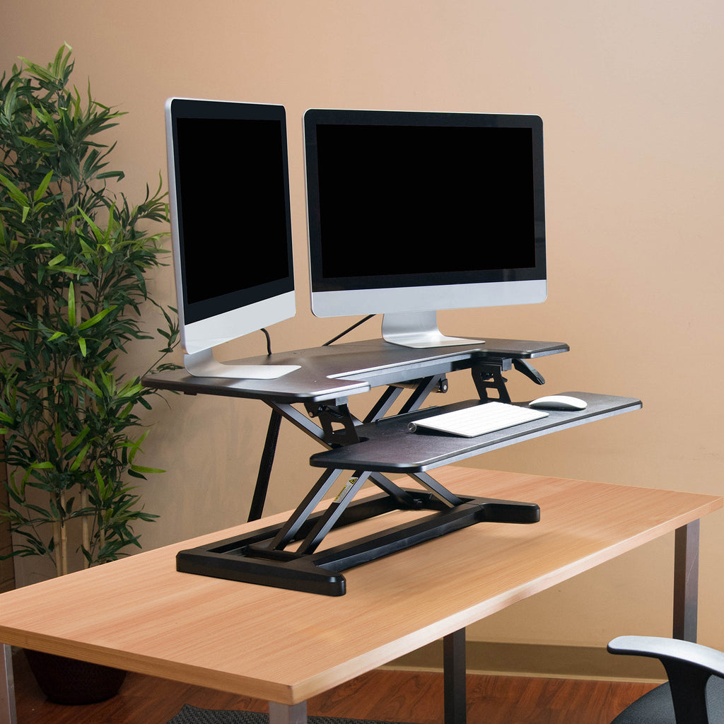 Height-adjustable desk converter with dual monitors and keyboard tray on a wooden office desk, shown in a modern workspace.