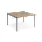 Two-person back-to-back office desk with oak-effect worktops and silver metal legs, shown against a white background.