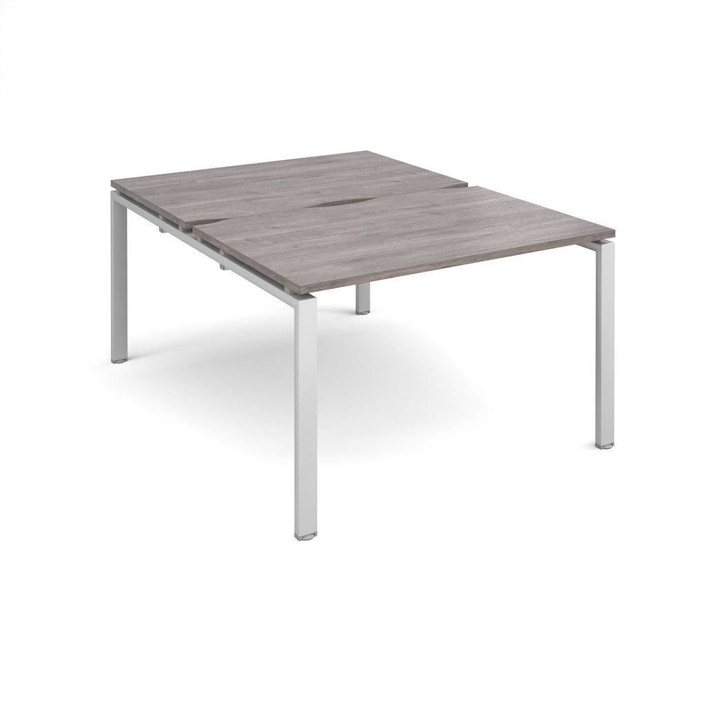 Large modular bench desk with a grey wood-effect top, integrated cable access cut-out, and silver metal legs, shown on a white background.
