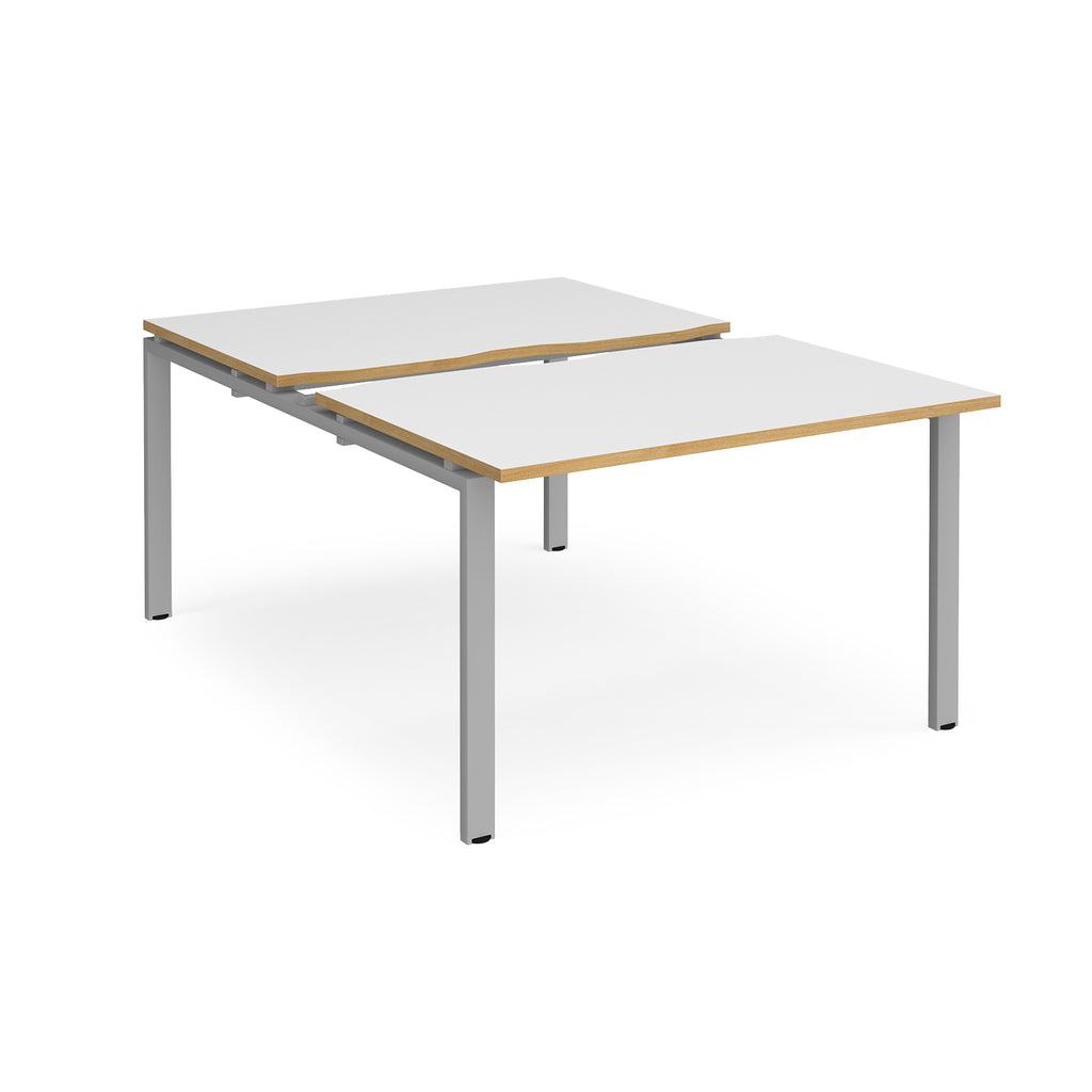 Large modular bench desk with white panels and wood-effect edging, integrated cable access cut-outs, and silver metal legs, shown on a white background.
