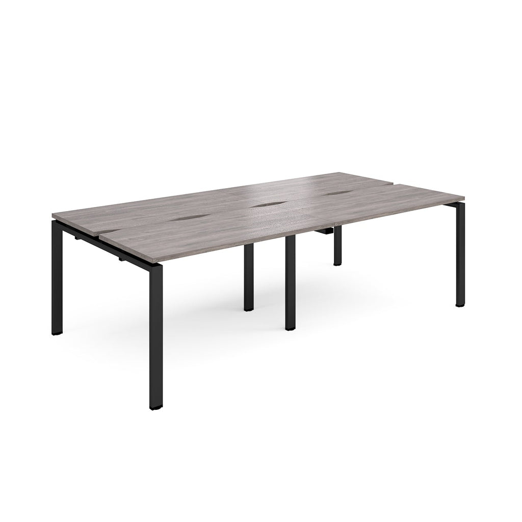 Large modular bench desk with a grey wood-effect top, integrated cable access cut-outs, and black metal legs, shown on a white background.