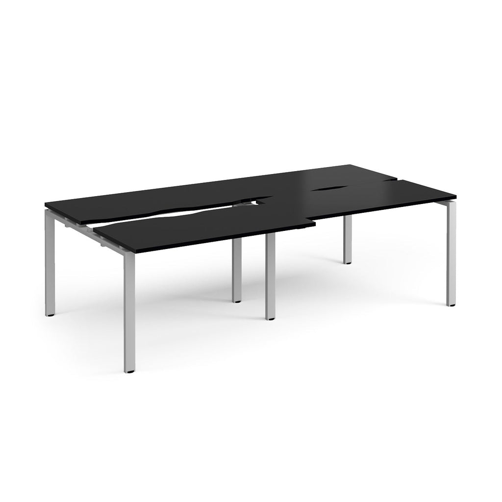 Large modular bench desk with a black worktop, integrated cable access cut-outs, and matching silver metal legs, shown on a white background.