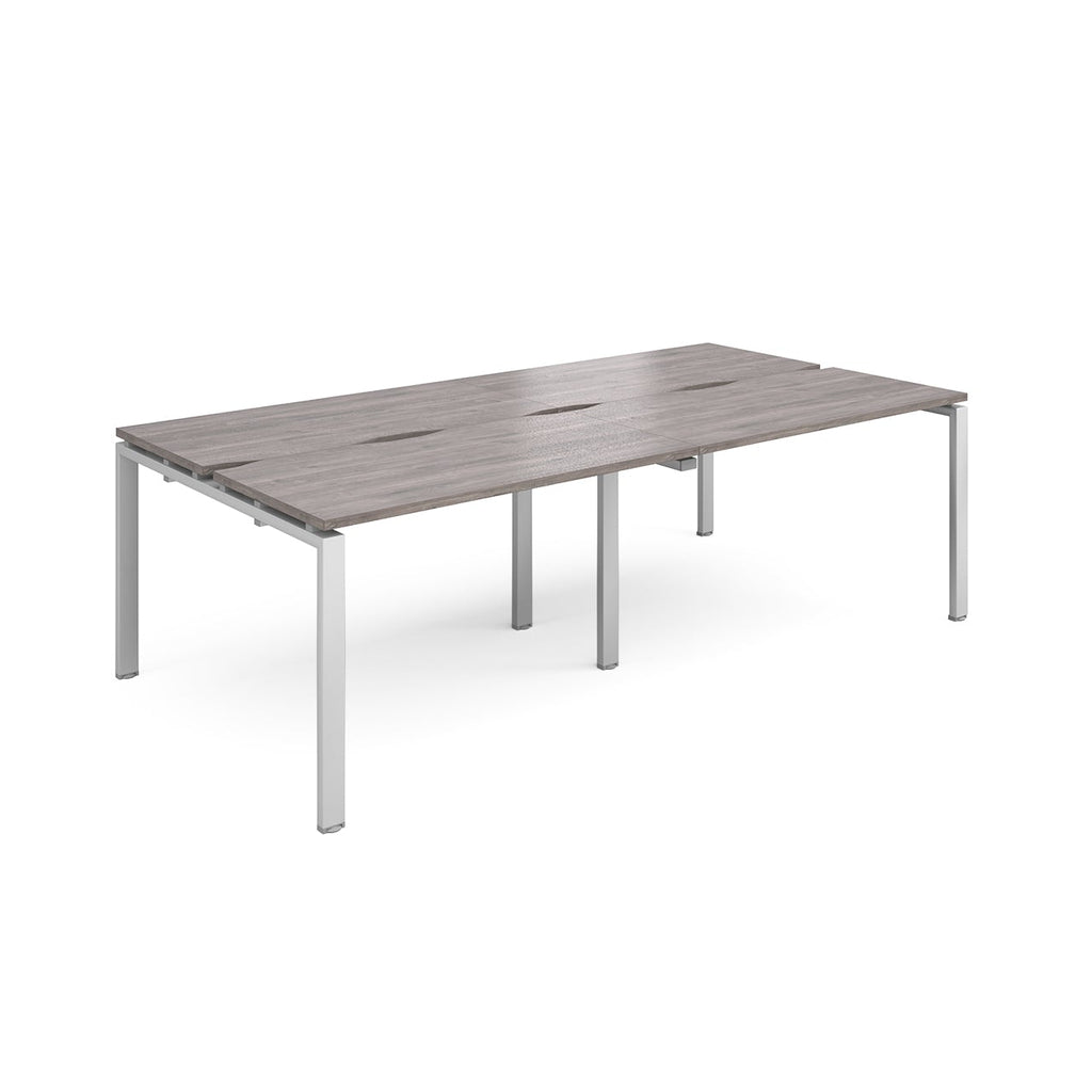 Large modular bench desk with a grey wood-effect top, integrated cable access slots, and silver metal legs, shown on a white background.