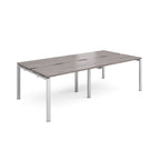 Large modular bench desk with a grey wood-effect top, integrated cable access slots, and silver metal legs, shown on a white background.