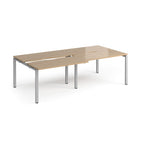 Large modular bench desk with a light oak wood-effect top, integrated cable access cut-outs, and silver metal legs, shown on a white background.