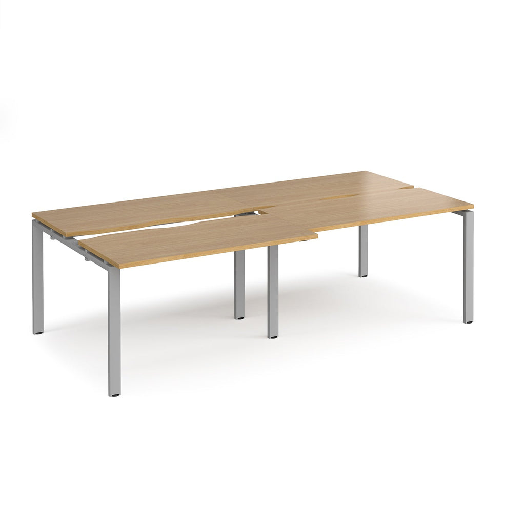 Large modular bench desk with a light oak wood-effect top, integrated cable access cut-outs, and silver metal legs, shown on a white background.