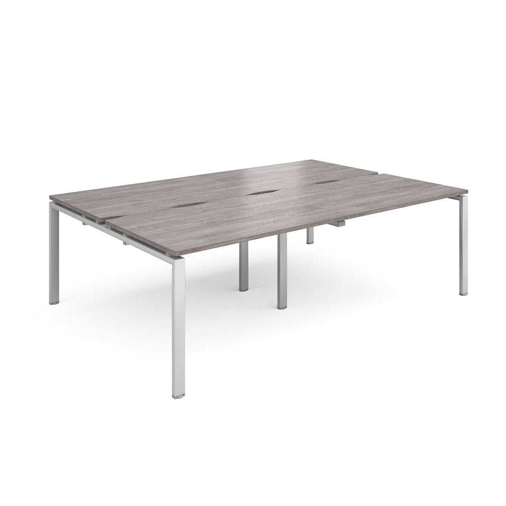 Large modular bench desk with a grey wood-effect top, integrated cable access slots, and silver metal legs, shown on a white background.