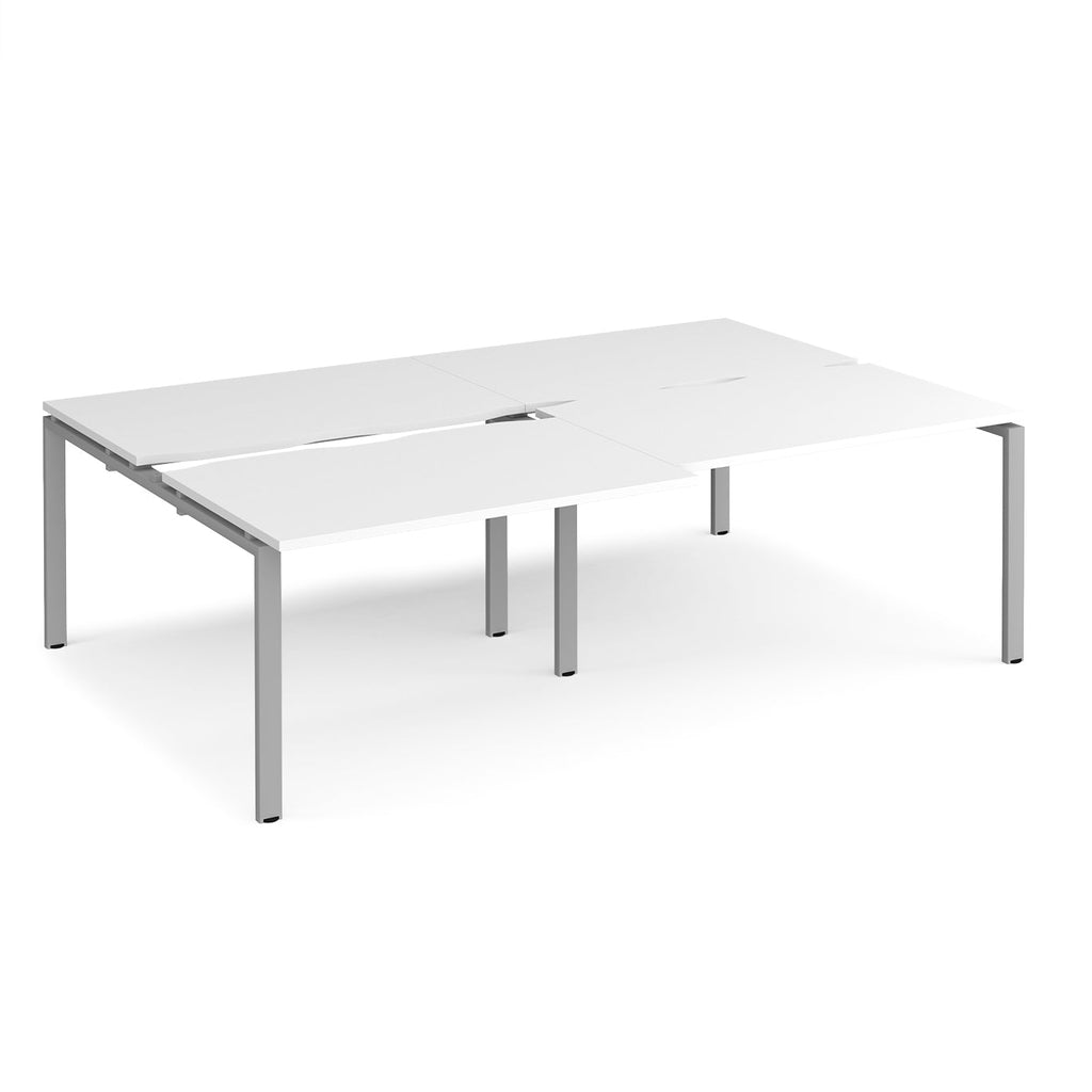 Large modular bench desk with a white worktop, integrated cable access cut-outs, and silver metal legs, shown on a white background.