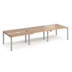 Large modular bench desk with a light wood-effect top, integrated cable cut-outs, and silver metal legs, shown on a white background.