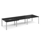 Large modular bench desk with a black worktop, integrated cable access cut-outs, and silver metal legs, shown on a white background.