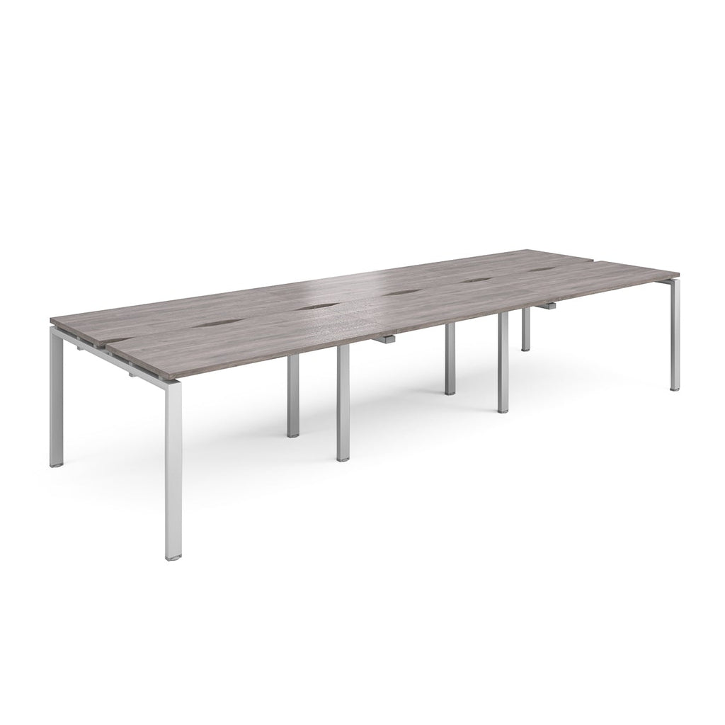 Large modular bench desk with a grey wood-effect top, integrated cable access slots, and silver metal legs, shown on a white background.