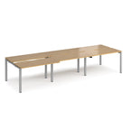 Large modular bench desk with a light wood-effect finish, integrated cable access cut-outs, and sturdy grey metal legs, shown on a white background.