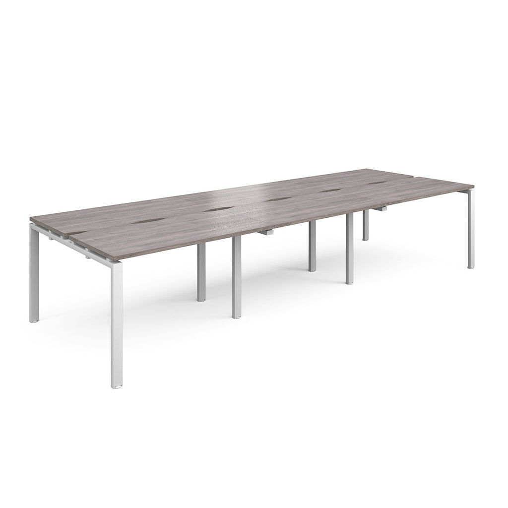 Large modular bench desk with a grey wood-effect top, integrated cable access slots, and white metal legs, shown on a white background.

