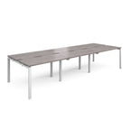 Large modular bench desk with a grey wood-effect top, integrated cable access slots, and white metal legs, shown on a white background.
