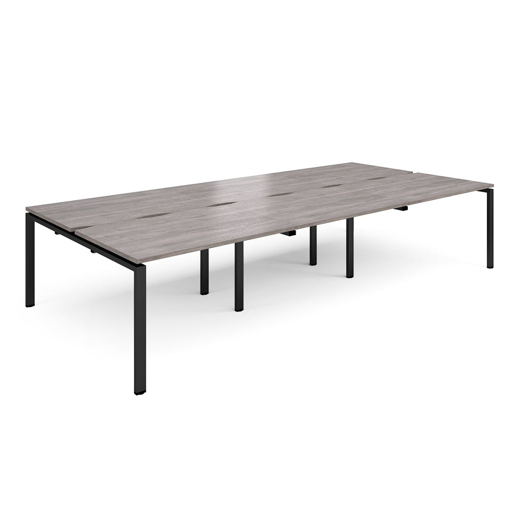 Large modular bench desk with a grey wood-effect top, integrated cable access slots, and black metal legs, shown on a white background.