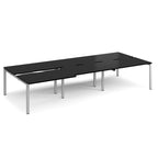 Large modular bench desk with a black worktop, integrated cable access cut-outs, and silver metal legs, shown on a white background.