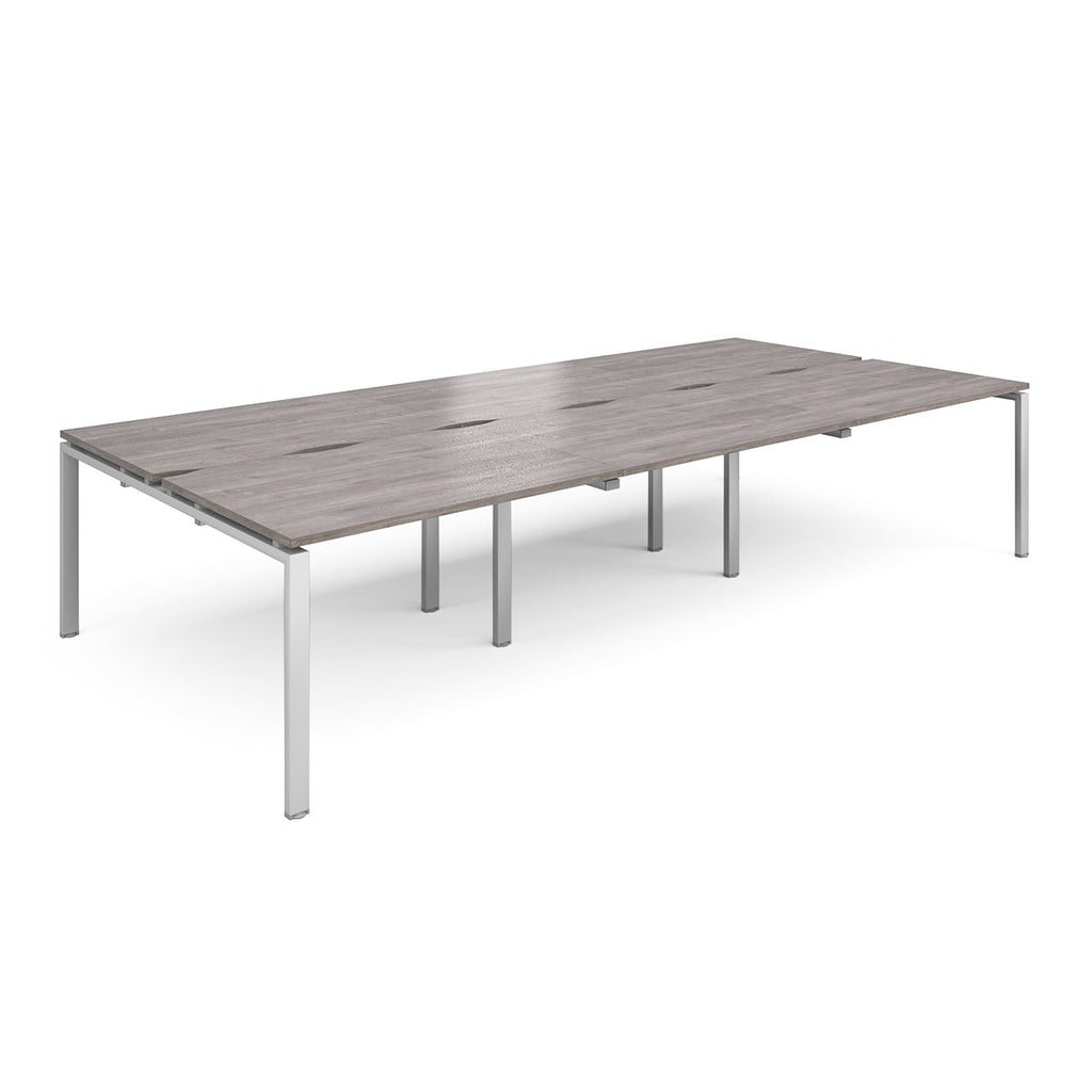 Large modular bench desk with a grey wood-effect top, integrated cable access slots, and silver metal legs, shown on a white background.
